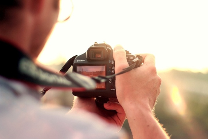 Symbolbild: Das Bild zeigt einen Mann mit einer Fotocamera in der Hand. Er ist von hinten zu sehen, wie er eine Landschaft fotografiert.