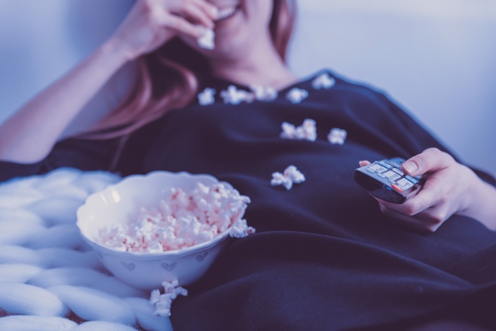 Symbolbild: Auf dem Bild sieht man eine junge Frau, die auf einem Bett liegt. Mit einer Hand schieb sie sich Popcorn in den Mund. In der anderen Hand hält sie eine Fernbedienung.