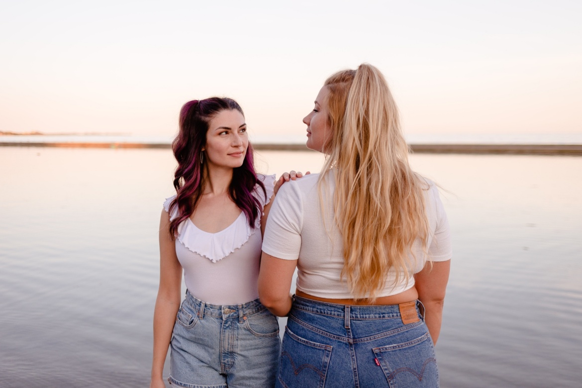 Zwei Freundinnen stehen eng zusammen an einem See und schauen sich an.