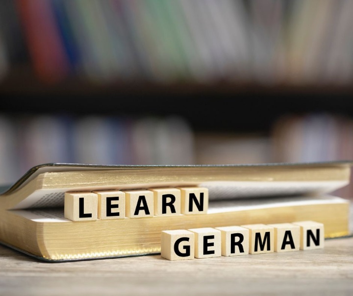 Ein Buch liegt auf einem Tisch. Die Seiten sind leicht aufgeklappt, dazwischen klemmen quadratische Holzklötze mit Buchstaben. Sie bilden die Aufforderung "Learn German" in Großbuchstaben.