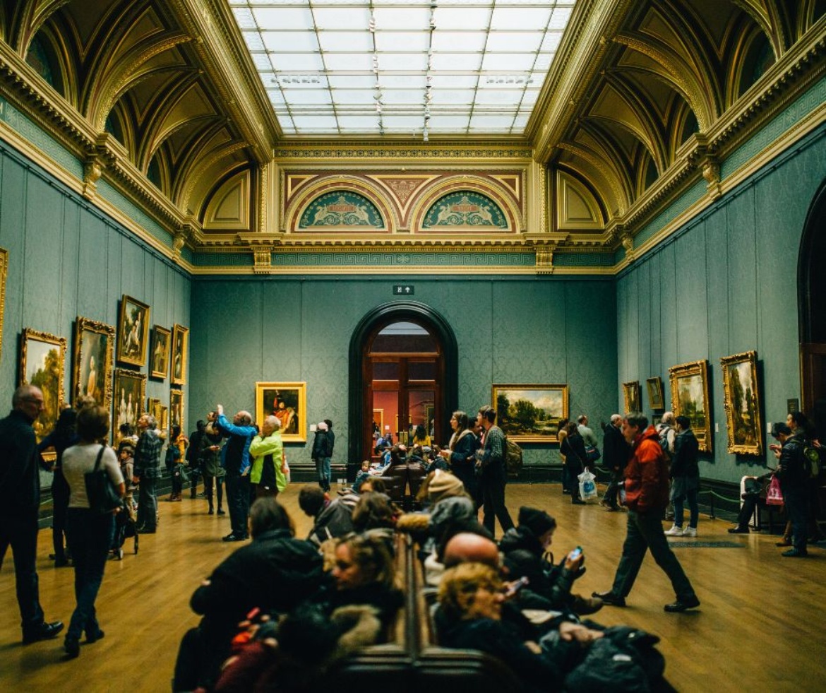 Ein Raum in einem Museum  Gemälde an den Wänden, eine Glaskuppel und Besucherinnen und Besucher gehen von Gemälde zu Gemälde.
