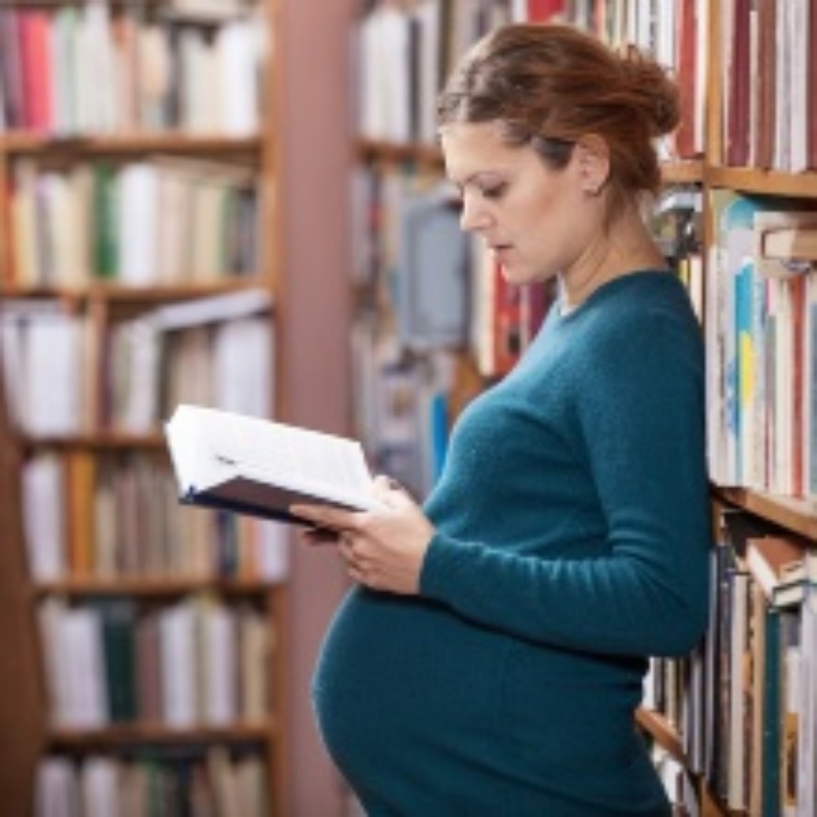 Eine schwangere Studentin lehnt an einem Bücherregal und liest ein Buch.