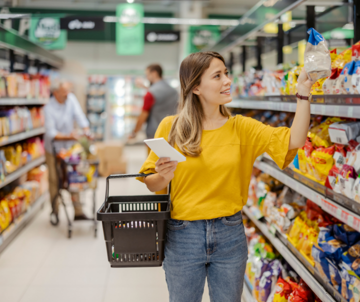 A young woman is walking through shelves in a supermarkt collecting groceries. 