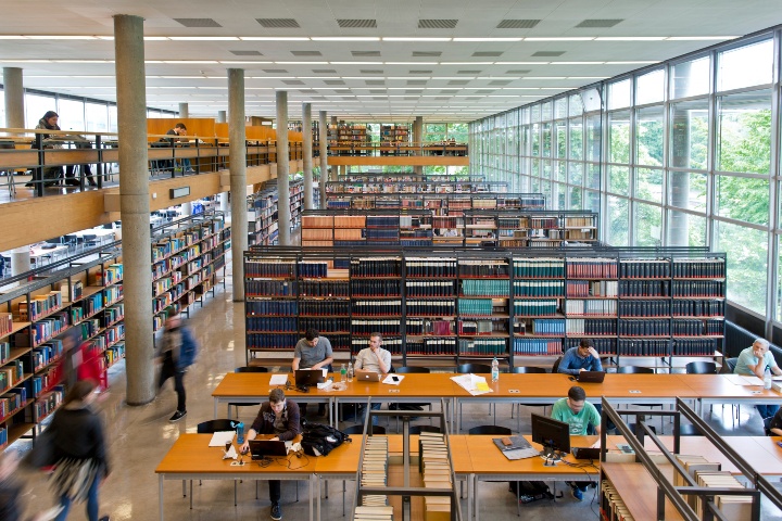 Von einer Erhähung aus in den Raum der Universitätsbibliothek hinunterfotografiert. Im Raum befinden isch Bücherregale und Lernplätze, an denen studierende lernen.