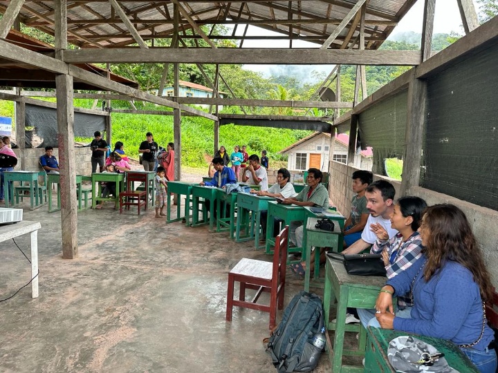 A group of people sits in a semicircle, each person at a table, in a wooden structure in the rainforest.