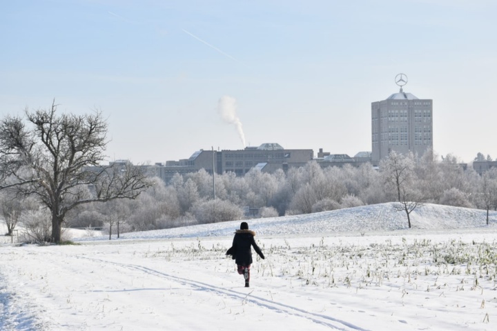 A snowy landscape, in the background trees, buildings and rising smoke from a chimney. A person walks through the winter landscape.