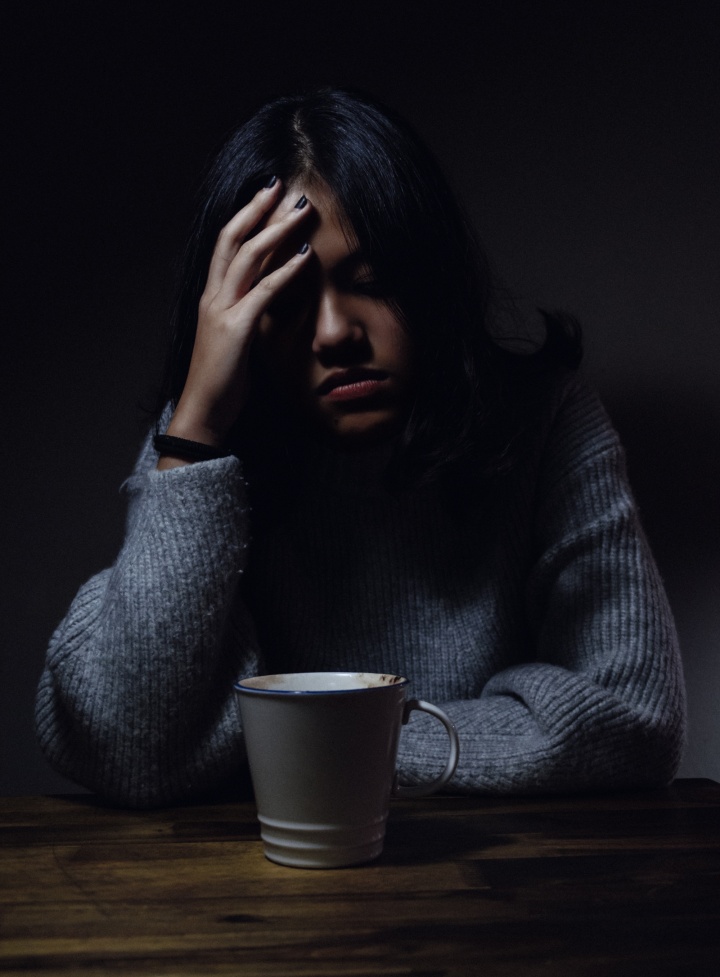 A young woman sits at a table in a dark room. She apears depressed.