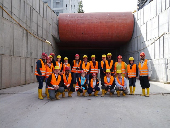 The picture shows a group of people. They are wearing safety vests and helmets and are standing in front of a tunnel.
